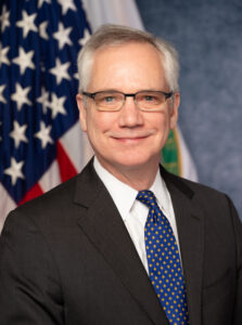 A white man wearing a suit and tie is smiling while seated in front of the American flag.