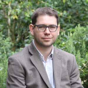 White man with short brown hair and black rimmed glasses dressed in a gray-brown suit and button down shirt with greenery in the background